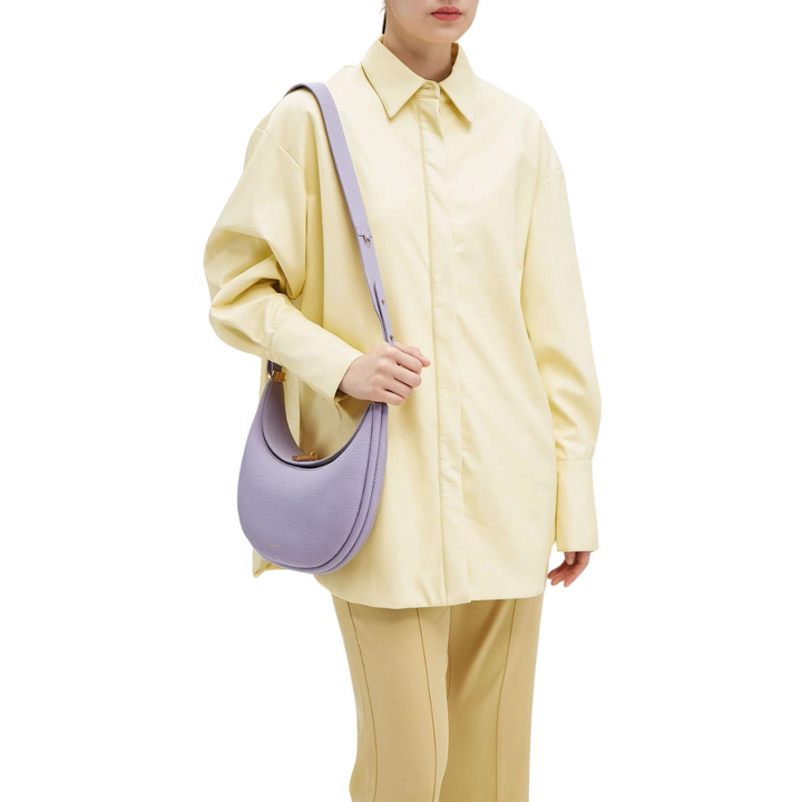 Una persona lleva una camisa amarilla de manga larga y pantalones beige, sosteniendo un bolso morado claro en su hombro. El fondo es blanco.