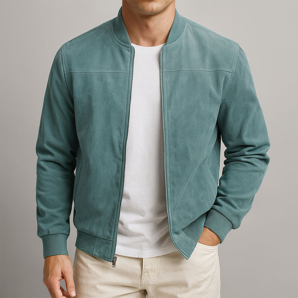 Una persona con una chaqueta bomber de color verde azulado claro sobre una camiseta blanca, con las manos delante. El fondo es liso y neutro.