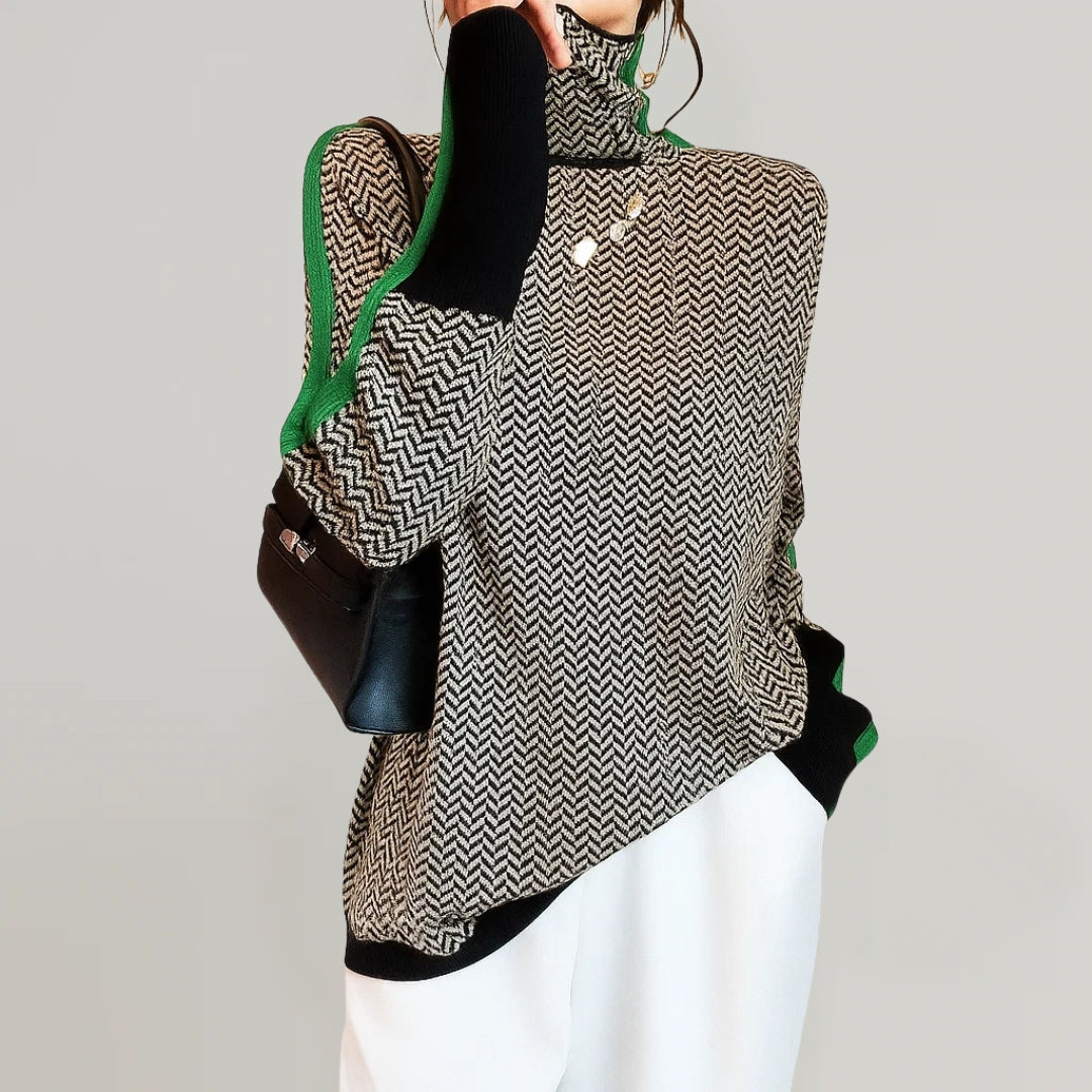 Un suéter de cuello alto con un diseño de patrón de zigzag en tonos gris y negro, con detalles en verde. La modelo lo combina con pantalones blancos y sostiene un bolso negro.